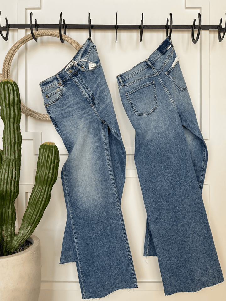 Two pairs of The Aiden High Rise Wide Leg Jeans by Savannah Sevens western life{&}style hang side by side on black hooks against a white paneled wall, with a potted cactus on the wooden floor to the left.