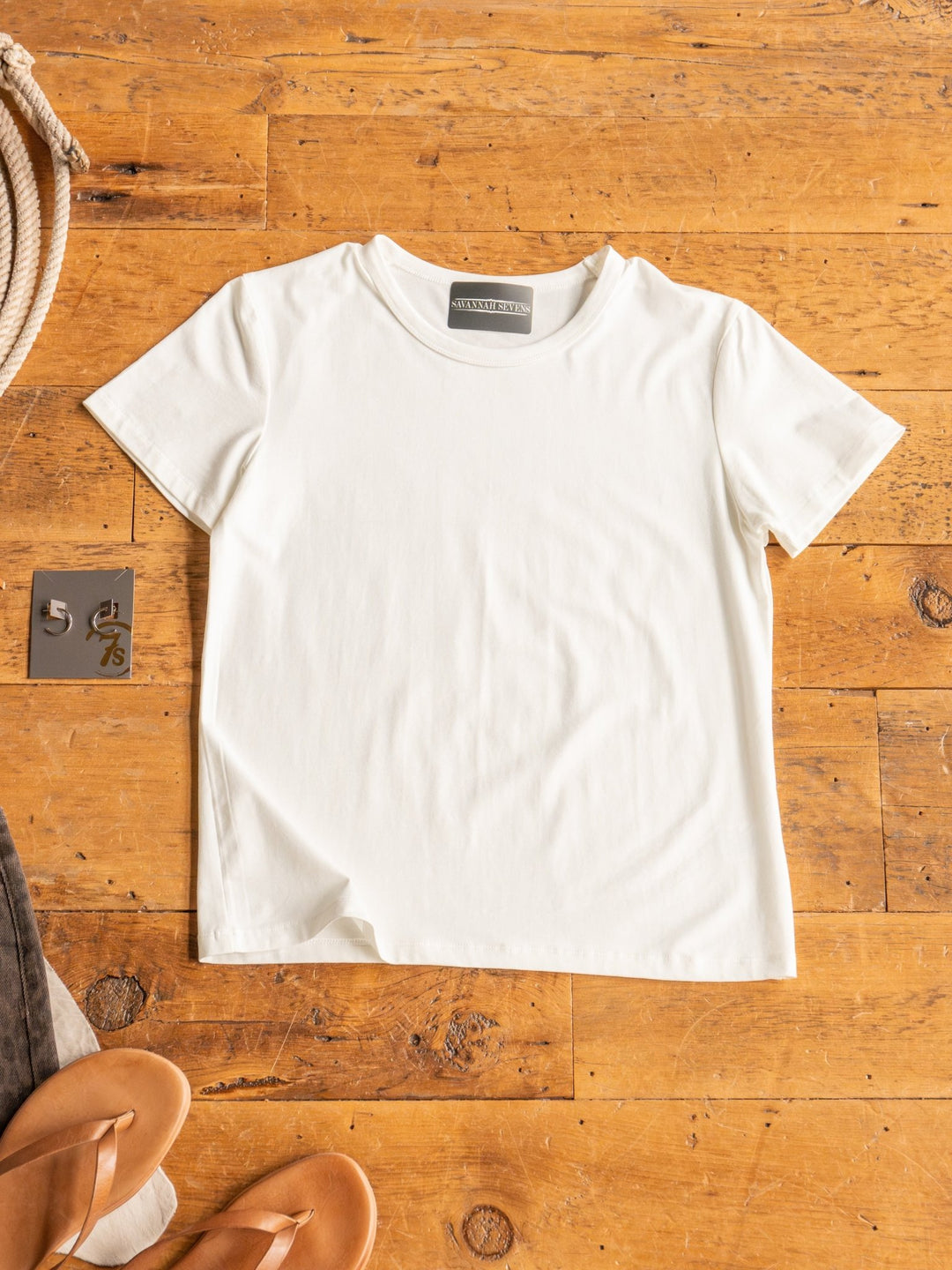 The Ailey by Savannah Sevens western life{&}style, a semi-fitted crew neck white short-sleeve top made from stretchy cotton, is laid flat on a wooden floor beside tan sandals and a small metal number tile for a versatile look.