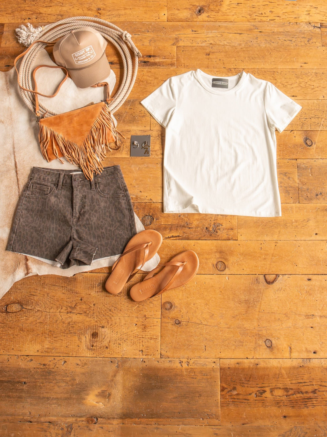 Flat lay of summer outfit featuring The Ailey by Savannah Sevens western life{&}style: semi-fitted crew neck cotton top, brown leopard print shorts, tan flip-flops, fringed shoulder bag, tan cap, and coiled rope on a wooden floor.
