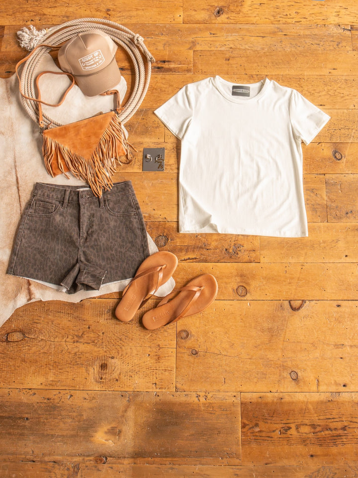 Flat lay of summer outfit featuring The Ailey by Savannah Sevens western life{&}style: semi-fitted crew neck cotton top, brown leopard print shorts, tan flip-flops, fringed shoulder bag, tan cap, and coiled rope on a wooden floor.