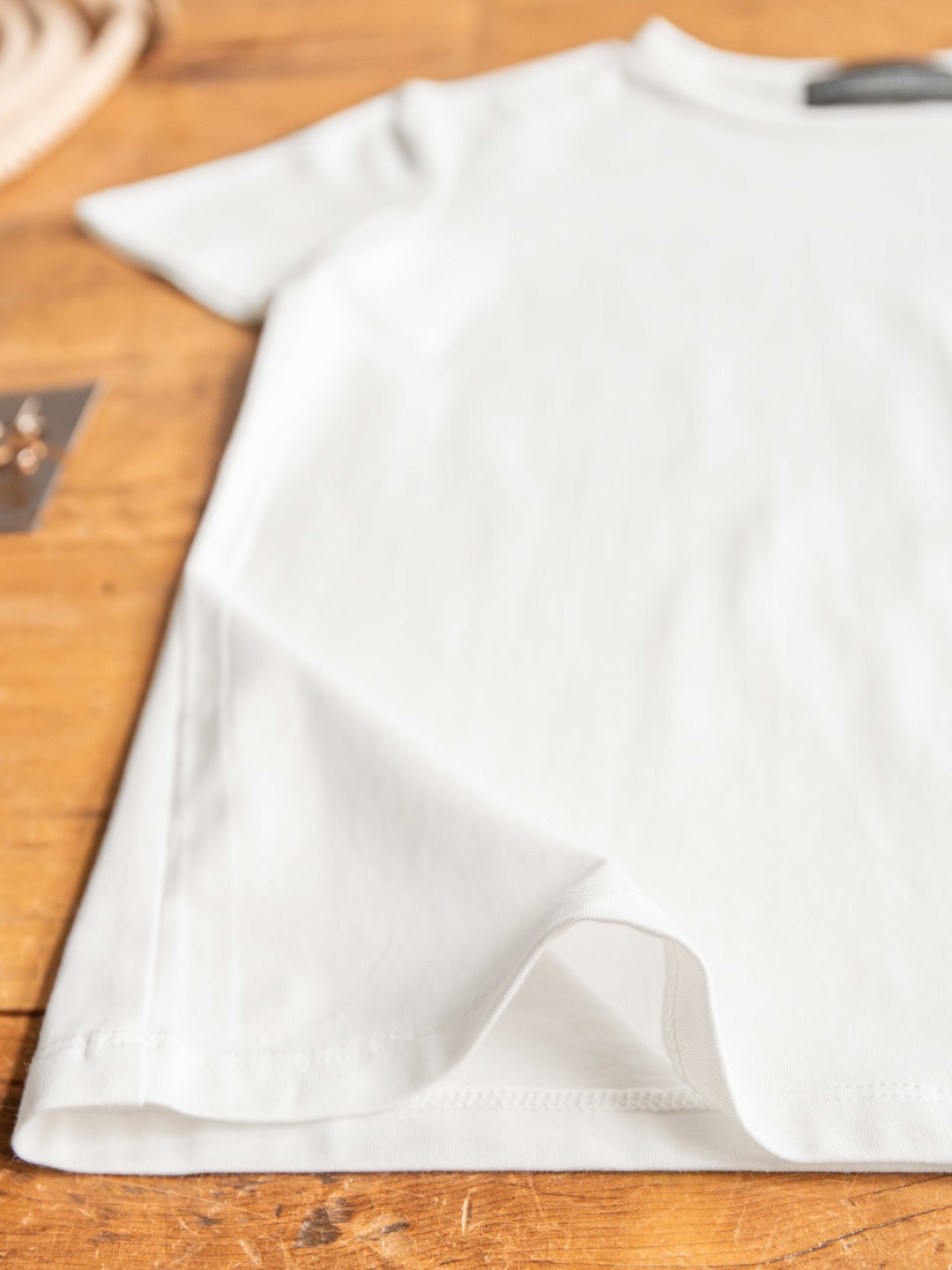A close-up of The Ailey, a semi-fitted white crew neck short sleeve top by Savannah Sevens western life{&}style, neatly laid on a wooden surface with part of the hem slightly lifted and blurred objects in the background.