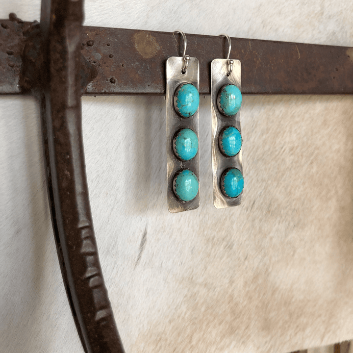 A pair of Alamosa Earrings by Savannah Sevens western life{&}style, featuring three round Kingman turquoise stones in each rectangular handmade piece, displayed against a soft white and brown background on a rustic metal rod.