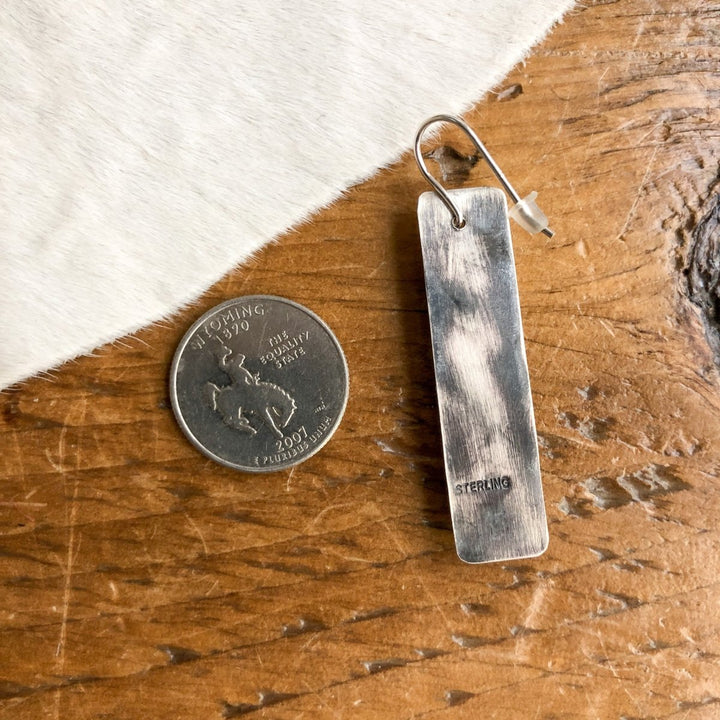 A  U.S. quarter is placed next to the Alamosa Earrings by Savannah Sevens western life{&}style, showcasing their rectangular handmade sterling silver design with hooks on a wooden surface beside white fur.
