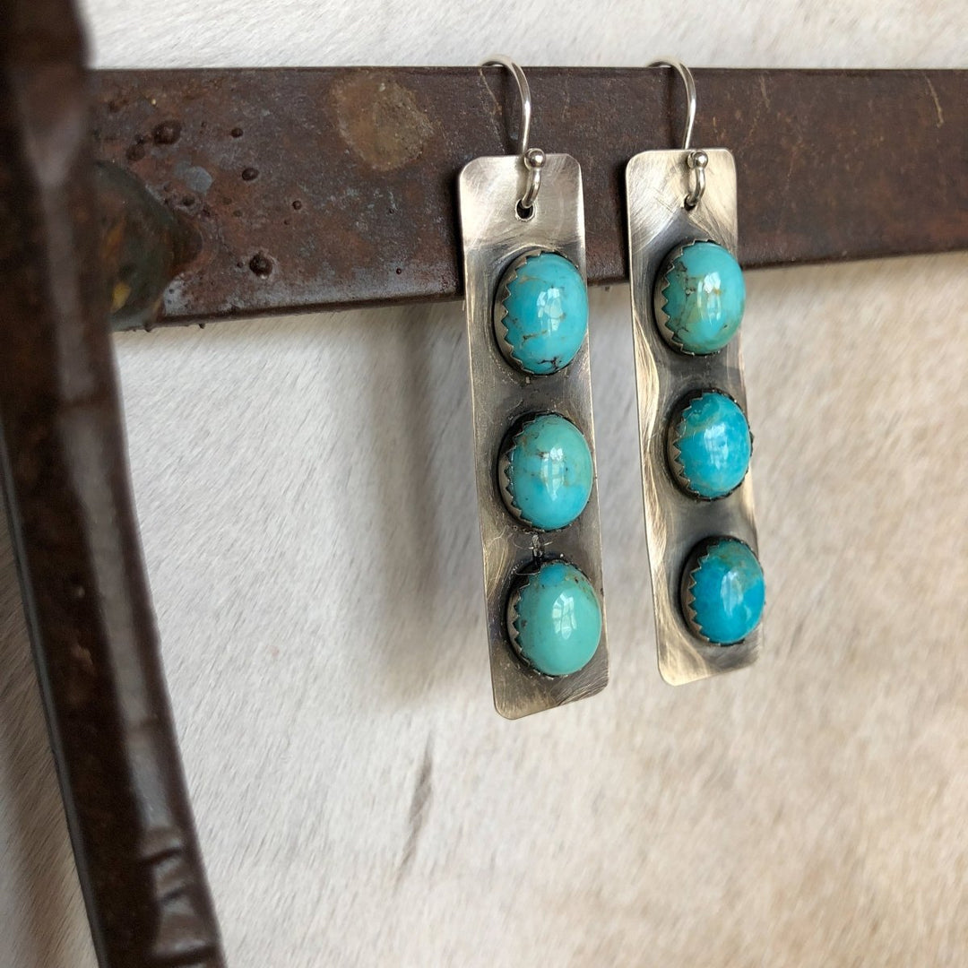 The Alamosa Earrings by Savannah Sevens western life{&}style feature three round Kingman turquoise stones set in rectangular sterling silver, hanging from a rustic metal bar against a light background.
