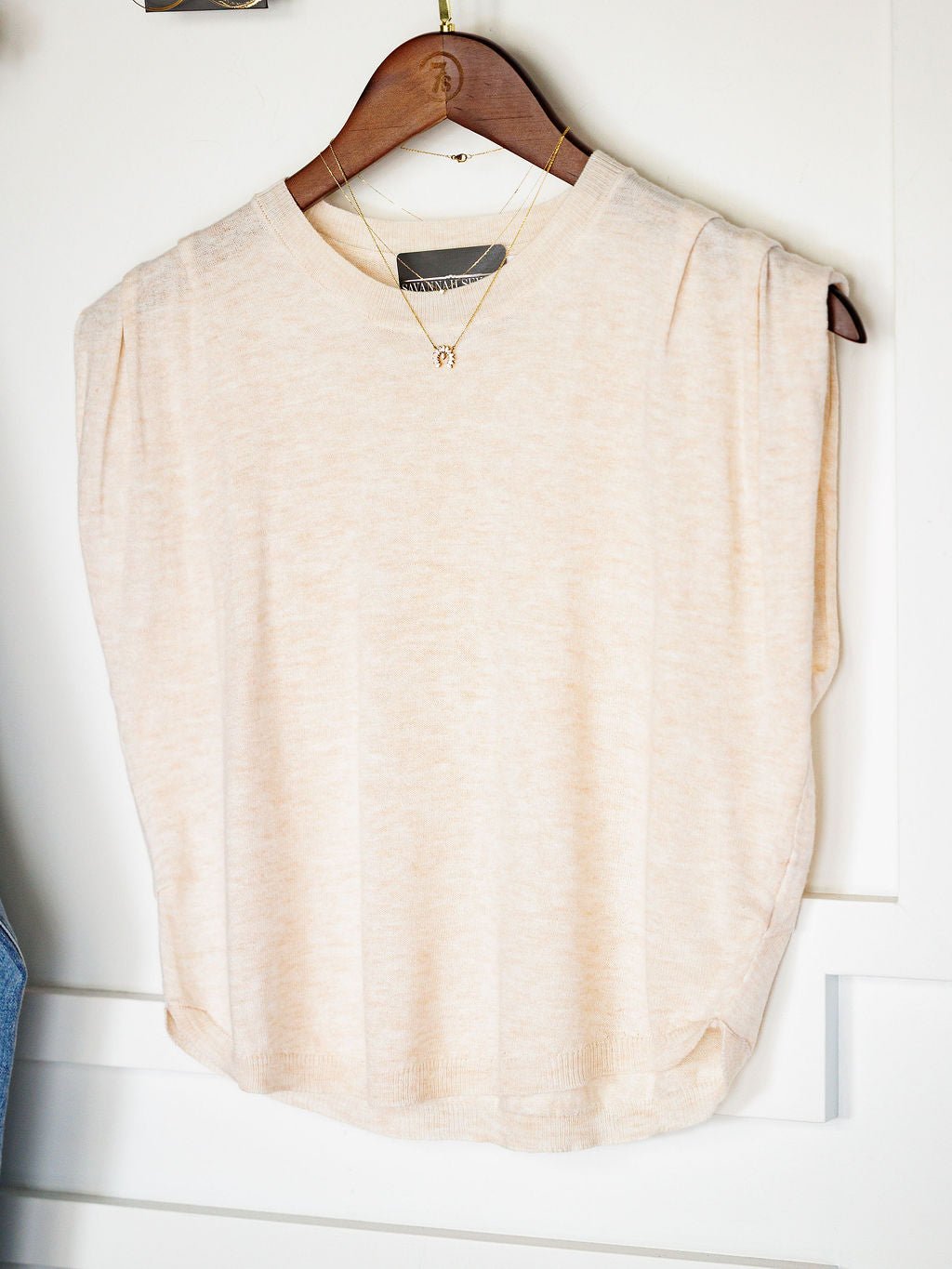 The Alcoa Oatmeal Wool Blend Top by Savannah Sevens western life{&}style, a relaxed-fit, sleeveless top, is displayed on a wooden hanger with a gold necklace over the neck, hanging on a white door.