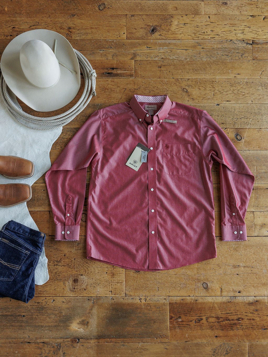 The Alden Men's Rhubarb Ariat Button Up by Savannah Sevens western life{&}style is displayed with tags on a wooden floor beside a white cowboy hat, brown boots, and folded blue jeans.