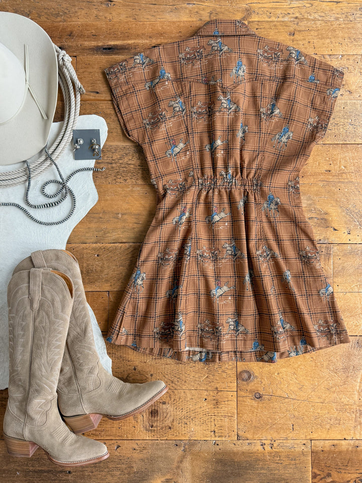 The Savannah Sevens western life{&}style Aledo Western Plaid Pearl Snap Dress, featuring brown plaid with blue floral accents, is displayed on a wooden floor alongside tan cowboy boots, a white cowboy hat, coiled rope, and a silver belt buckle.