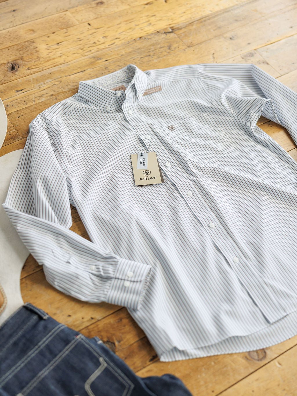 The Allendale Ariat Men's Striped Button Up by Savannah Sevens western life{&}style, in light blue and white with tags, is laid flat on a wooden floor next to dark blue jeans.