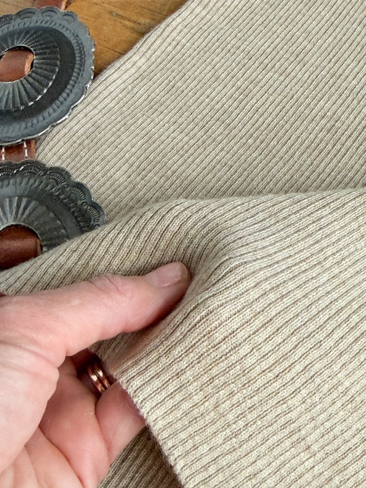 A hand holds the edge of the Almont Oatmeal Knit Top by Savannah Sevens western life{&}style. Metal pastry cutters with fluted edges are partially visible in the background on a wooden surface.
