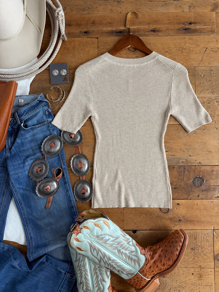 The Almont Oatmeal Knit Top from Savannah Sevens western life{&}style with short sleeves hangs alongside blue jeans, turquoise cowboy boots, a brown belt, a silver concho belt, earrings, a white rope, and a white hat—all neatly arranged on a wooden floor.