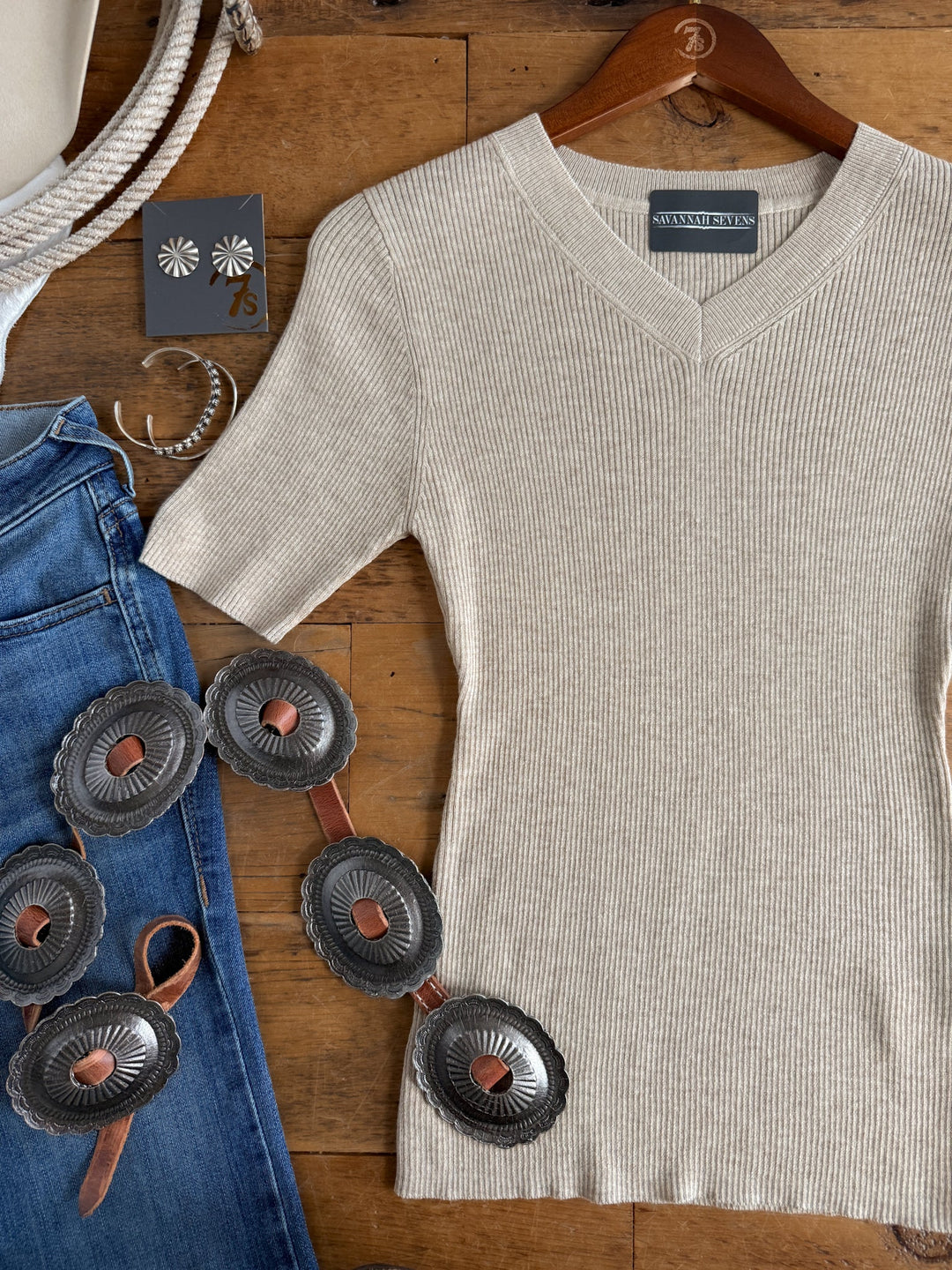 The Almont Oatmeal Knit Top by Savannah Sevens western life{&}style is laid on a wooden surface next to blue jeans, a silver concho belt, silver hoop earrings, and daisy-shaped silver stud earrings.