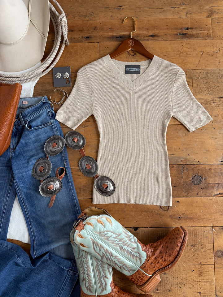 Flat lay of the Savannah Sevens western life{&}style Almont Oatmeal Knit Top on a hanger, paired with blue jeans, turquoise and brown cowboy boots, a leather concho belt, a brown purse, a white cowboy hat, rope, and black daisy earrings displayed on a wooden floor.