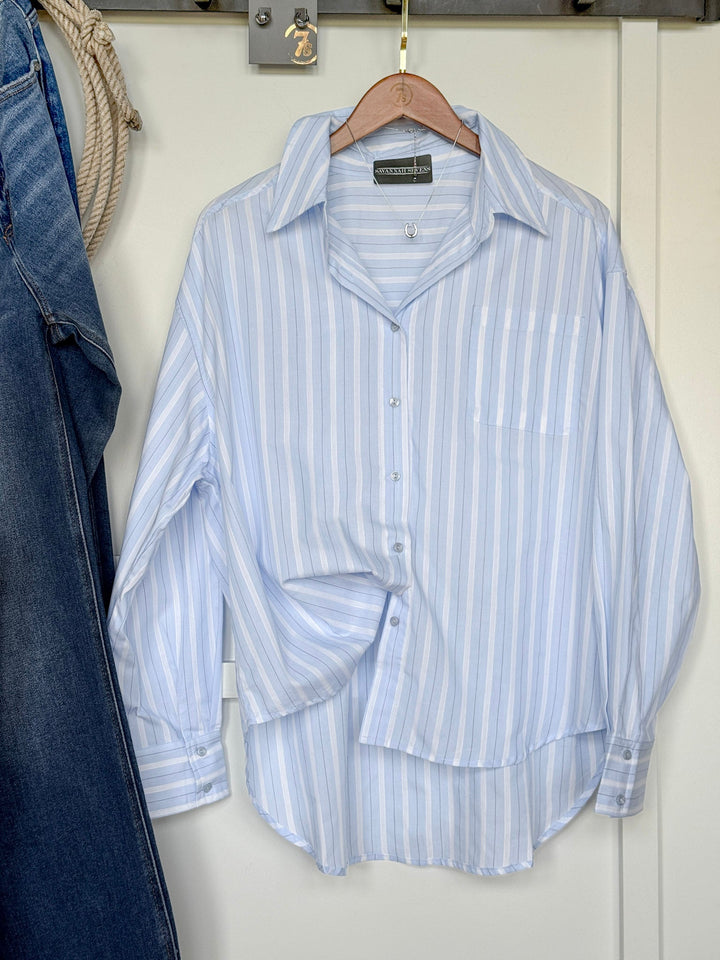 The Altavista Striped Button Up Oversized Top by Savannah Sevens western life{&}style, featuring light blue and white stripes and long sleeves, hangs on a wooden hanger against a white door, with a pair of blue jeans visible to the left.