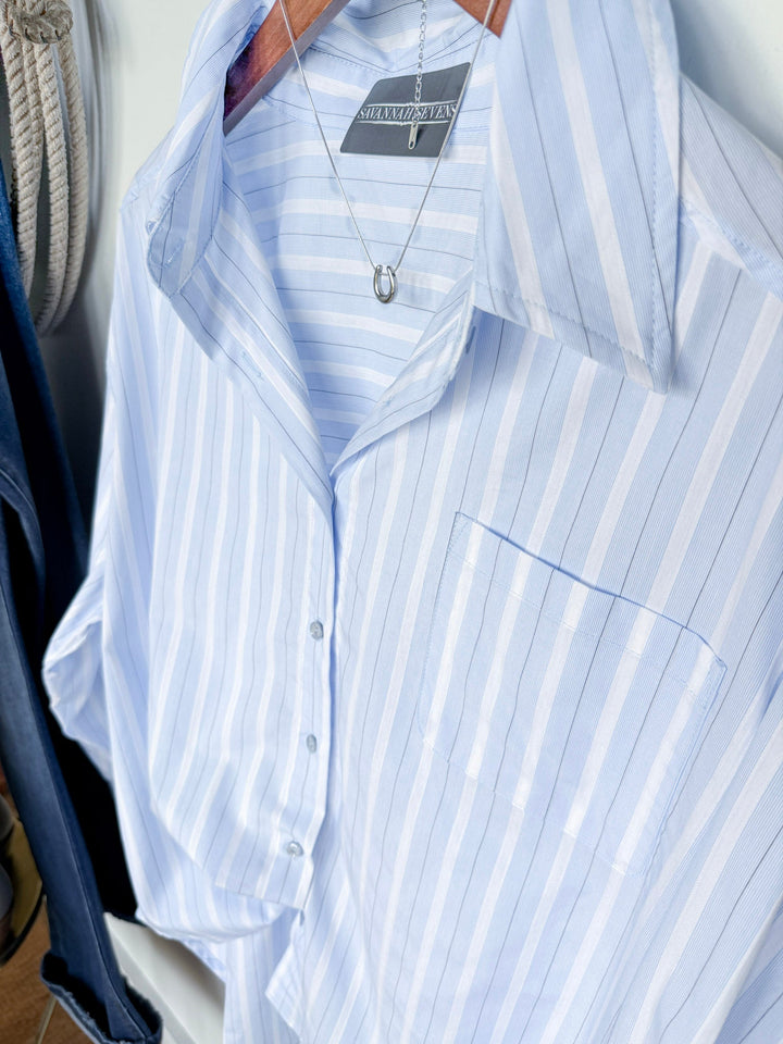 The Altavista Striped Button Up Oversized Top by Savannah Sevens western life{&}style, featuring light blue and white stripes, hangs on a wooden hanger, with a silver horseshoe pendant necklace draped around its collar.