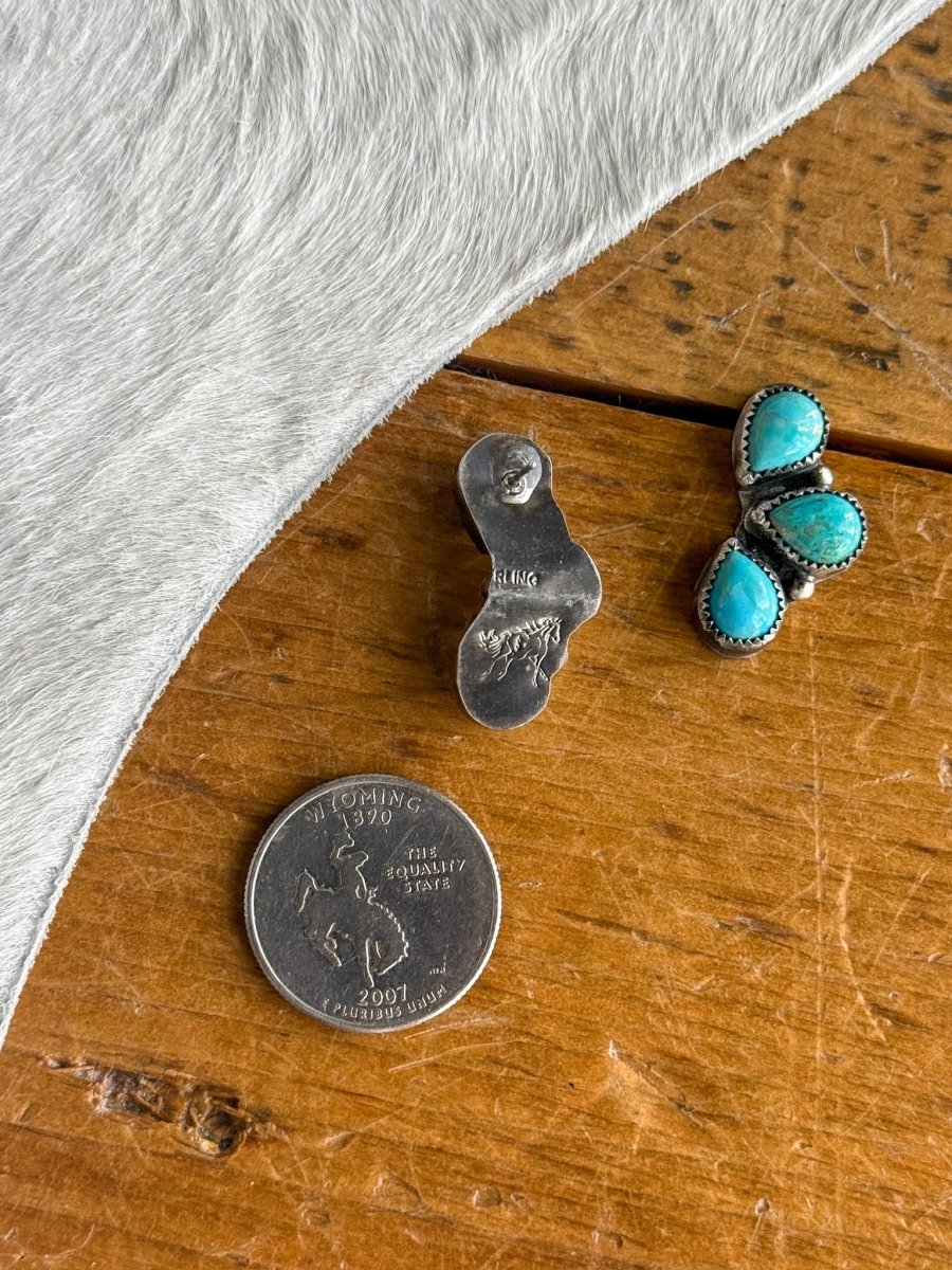 The Amargosa Earrings from Savannah Sevens western life{&}style, featuring turquoise and silver with a sterling stamp and engraved horse on the back, rest beside a Wyoming quarter on wood accented with light cowhide—showcasing fine craftsmanship.