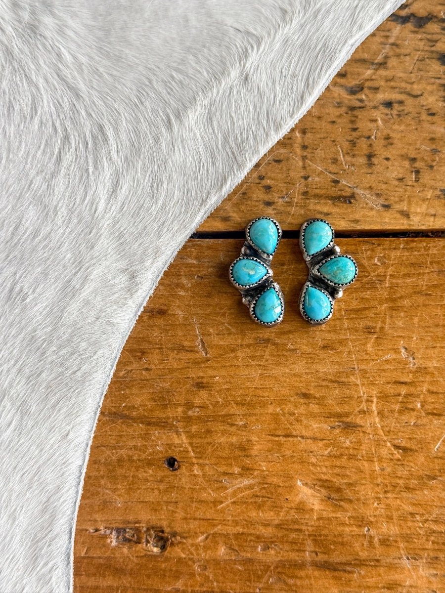 The Amargosa Earrings by Savannah Sevens western life{&}style feature three turquoise stones in a triangular pattern set in sterling silver, displayed on a wooden surface beside light-colored cowhide.
