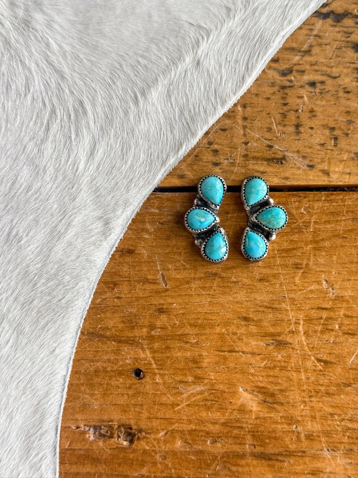 The Amargosa Earrings by Savannah Sevens western life{&}style feature three turquoise stones in a triangular pattern set in sterling silver, displayed on a wooden surface beside light-colored cowhide.