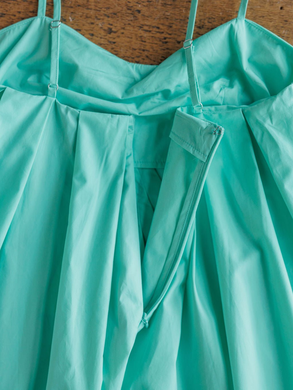 A close-up of the Amasa Seafoam Cotton Dress by Savannah Sevens western life{&}style, showing sleeveless design, petite adjustable straps, pleats, and a partially unzipped side zipper against a wooden background.