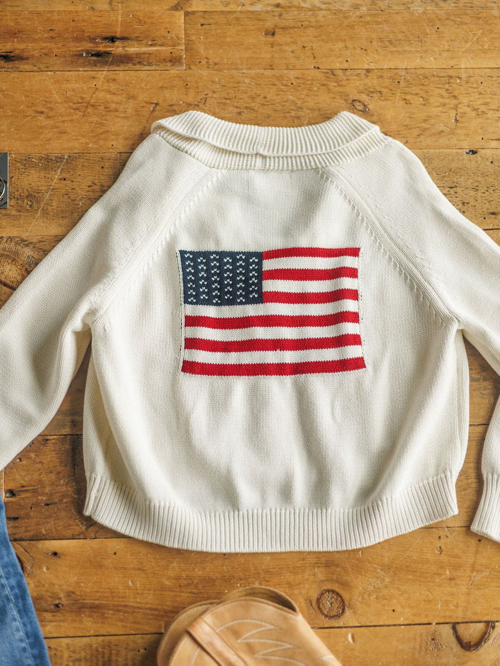 The American Classic {Exclusive} by Savannah Sevens western life{&}style is a white 100% cotton cardigan featuring an American flag design on the back, displayed on wood beside a sandal and blue denim—an exclusive Savannah Sevens piece.