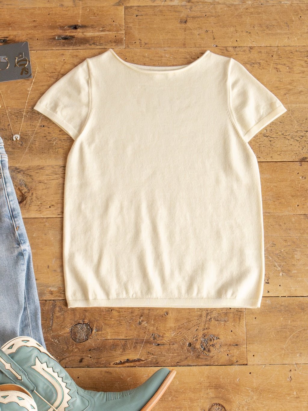 The Amery Buttercream Knit Top by Savannah Sevens western life{&}style, crafted from soft quality fabric, is displayed flat on a wooden floor beside the edge of blue jeans, a necklace, and green and beige cowboy boots. Its ribbed short sleeves provide subtle texture to this timeless piece.