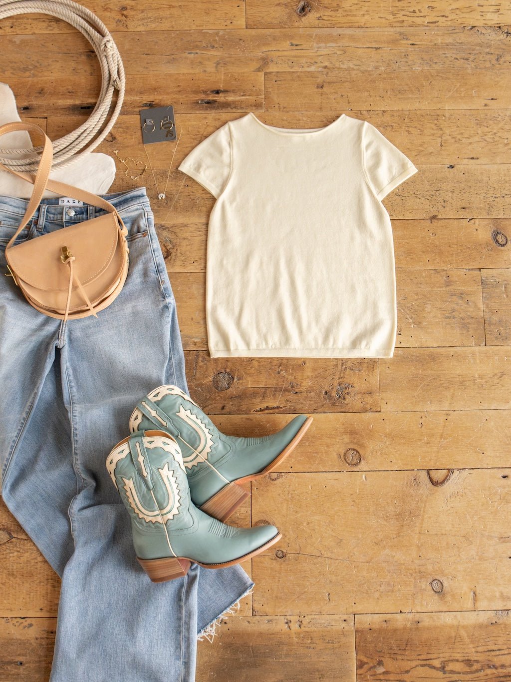 Styled in a Western theme on a wooden floor: light blue jeans, the Amery Buttercream Knit Top by Savannah Sevens western life{&}style featuring ribbed short sleeves, a tan crossbody bag, turquoise cowboy boots with white trim, wide-brim hat, and silver earrings. The soft quality fabric of the Amery top brings both comfort and style to this classic outfit.