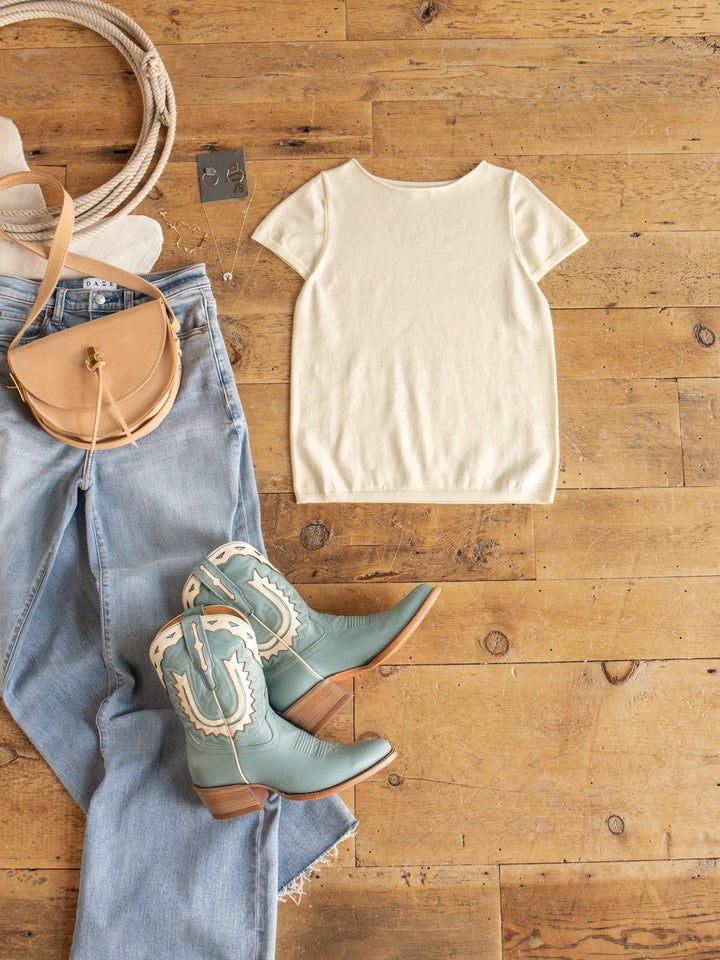 Styled in a Western theme on a wooden floor: light blue jeans, the Amery Buttercream Knit Top by Savannah Sevens western life{&}style featuring ribbed short sleeves, a tan crossbody bag, turquoise cowboy boots with white trim, wide-brim hat, and silver earrings. The soft quality fabric of the Amery top brings both comfort and style to this classic outfit.