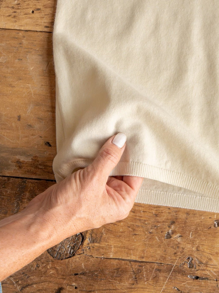 A hand with light-colored nail polish gently pinches the hem of the Amery Buttercream Knit Top by Savannah Sevens western life{&}style, made from soft quality fabric and resting on a rustic wooden surface. Ribbed sleeves add delicate detail to this beige knit top.
