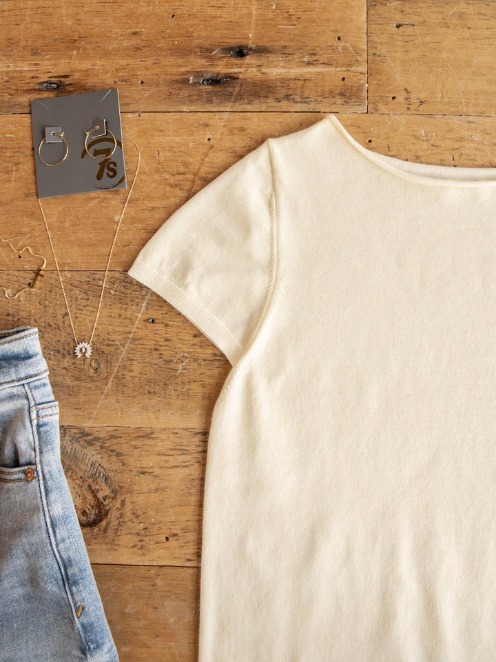 The Amery Buttercream Knit Top from Savannah Sevens western life{&}style is displayed beside light blue denim jeans, with a gold necklace featuring a small charm and a pair of gold earrings arranged on a wooden surface.