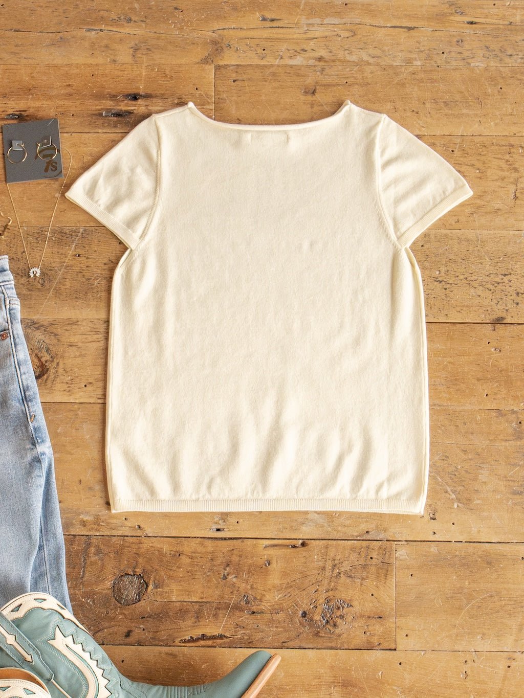 The Amery Buttercream Knit Top by Savannah Sevens western life{&}style is laid flat on a wooden floor. Blue jeans, green cowboy boots, a necklace, and a pair of earrings are arranged around the top at the edges of the image.