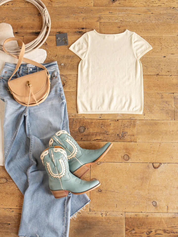 Flat lay of the Amery Buttercream Knit Top by Savannah Sevens western life{&}style with ribbed sleeves and soft quality fabric, paired with light blue cowboy boots, light-wash jeans, a tan crossbody bag, and jewelry on a wooden floor.
