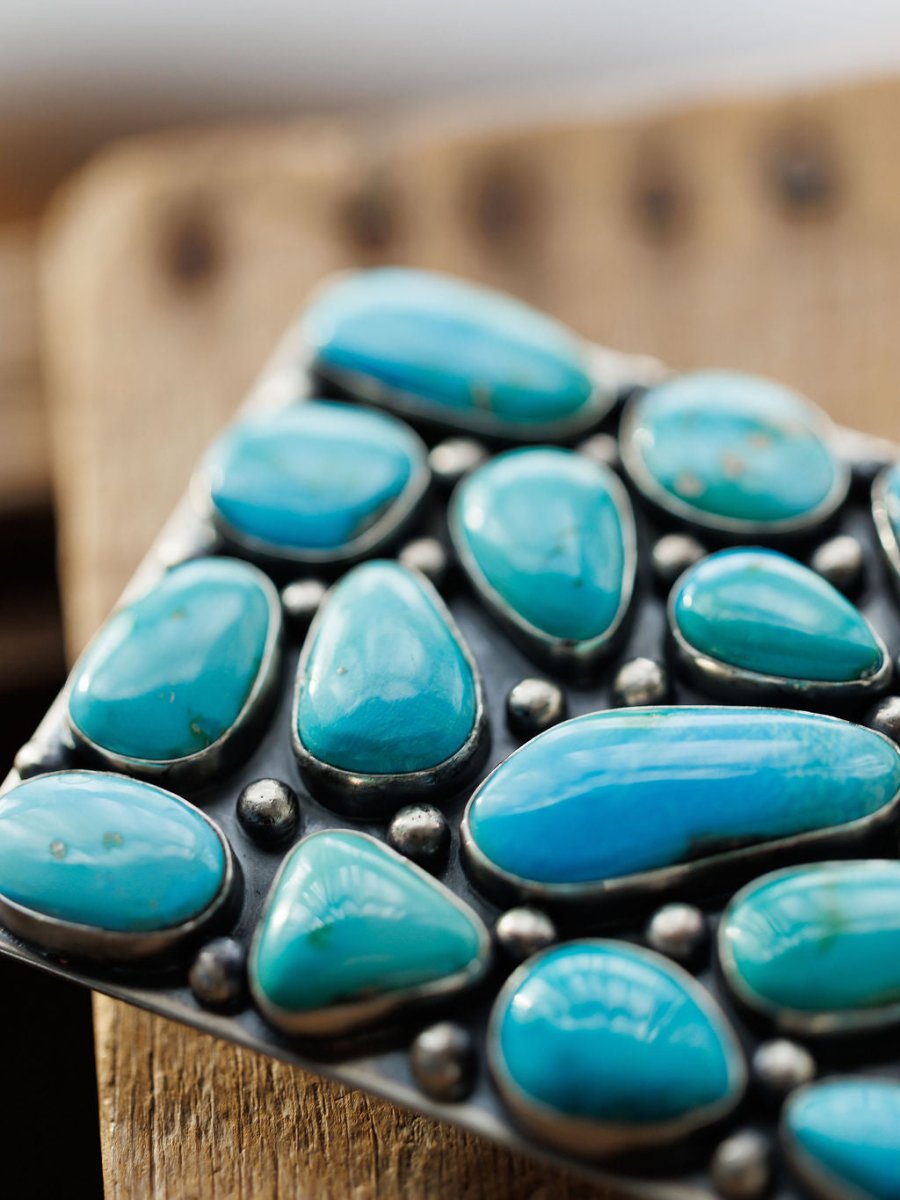 A close-up of the Amorita Buckle by Savannah Sevens western life{&}style, featuring polished turquoise stones set in silver on a rustic wooden surface.