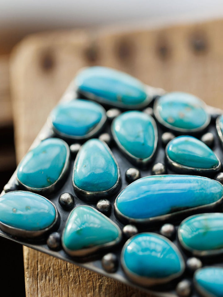 A close-up of the Amorita Buckle by Savannah Sevens western life{&}style, featuring polished turquoise stones set in silver on a rustic wooden surface.