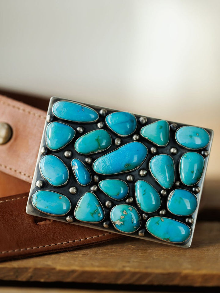 The Amorita Buckle by Savannah Sevens western life{&}style, a rectangular sterling silver piece with polished turquoise stones, is displayed on a brown leather belt atop a wooden surface.