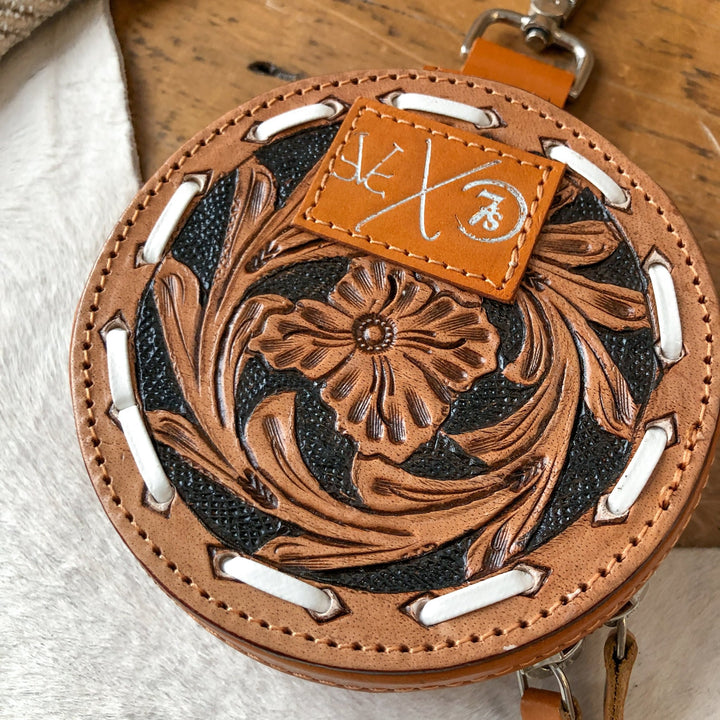 The Amorita Tooled Leather Card Holder by Savannah Sevens western life{&}style is a handmade, round leather case with a floral tooled design, black background, white lacing, and a tan patch with white markings shown on wood beside white fur.