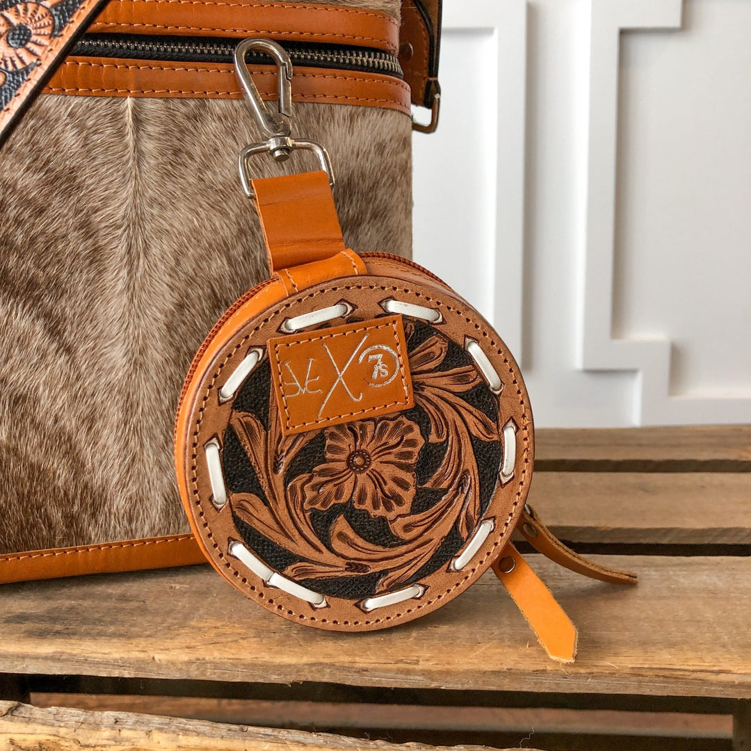 The Amorita Tooled Leather Card Holder by Savannah Sevens western life{&}style is a handmade round purse with floral patterns, white stitching, a zipper, and a leather patch with SLX 73, hanging from a brown and white cowhide bag.