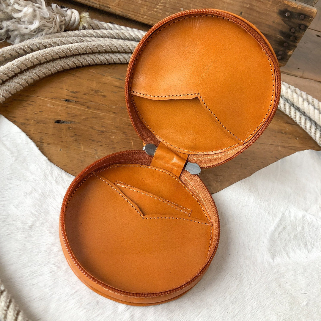 The Amorita Tooled Leather Card Holder by Savannah Sevens western life{&}style is a round, tan handmade leather case with a zipper and two interior pockets, displayed open on a textured surface near a coiled rope and wooden pieces.