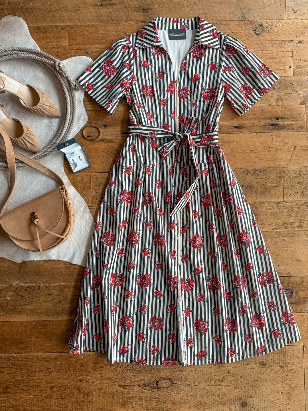 The Anaheim Striped Floral Dress by Savannah Sevens western life{&}style, featuring cream and black stripes with red floral patterns, is laid out on a wooden floor alongside a tan purse, beige heels, and a light hat.