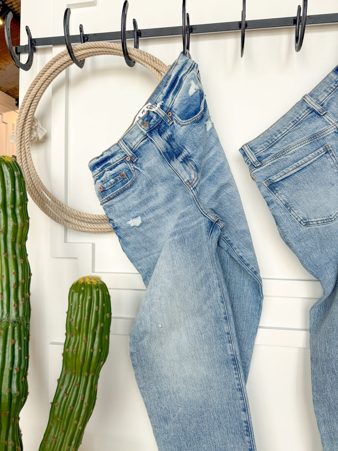 The Andie High Rise 90's Wide Leg Jean by Savannah Sevens western life{&}style hangs on a black metal rack near coiled rope and green cacti, set against a white paneled wall.