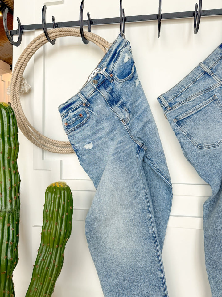 The Andie High Rise 90's Wide Leg Jean by Savannah Sevens western life{&}style hangs on a black metal rack near coiled rope and green cacti, set against a white paneled wall.