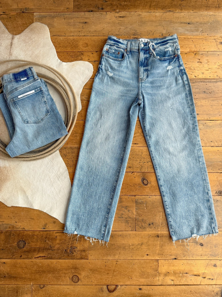 The Andie High Rise 90's Wide Leg Jean in light blue by Savannah Sevens western life{&}style lies flat with frayed hems next to a cowhide rug, beside a folded pair of vintage distressed jeans and a coiled rope.