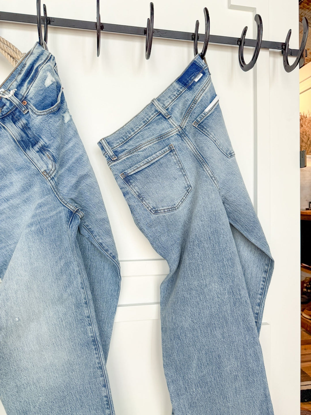 Two pairs of The Andie High Rise 90's Wide Leg Jeans by Savannah Sevens western life{&}style hang from black hooks on a white wall, showing back pockets and seams. A cozy interior glows softly in the blurred background.
