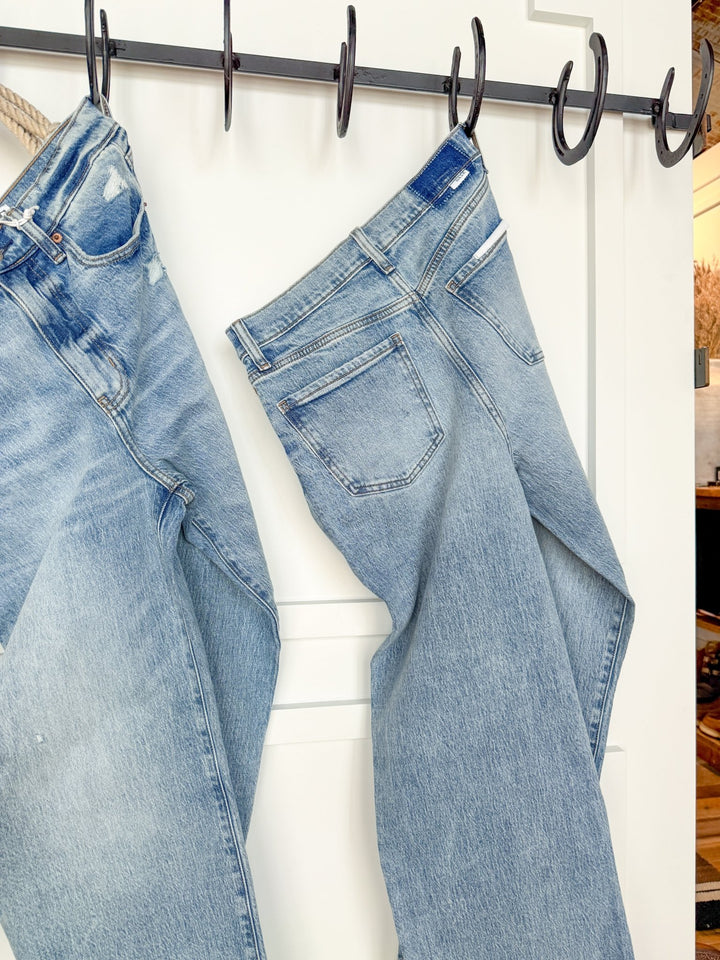 Two pairs of The Andie High Rise 90's Wide Leg Jeans by Savannah Sevens western life{&}style hang from black hooks on a white wall, showing back pockets and seams. A cozy interior glows softly in the blurred background.