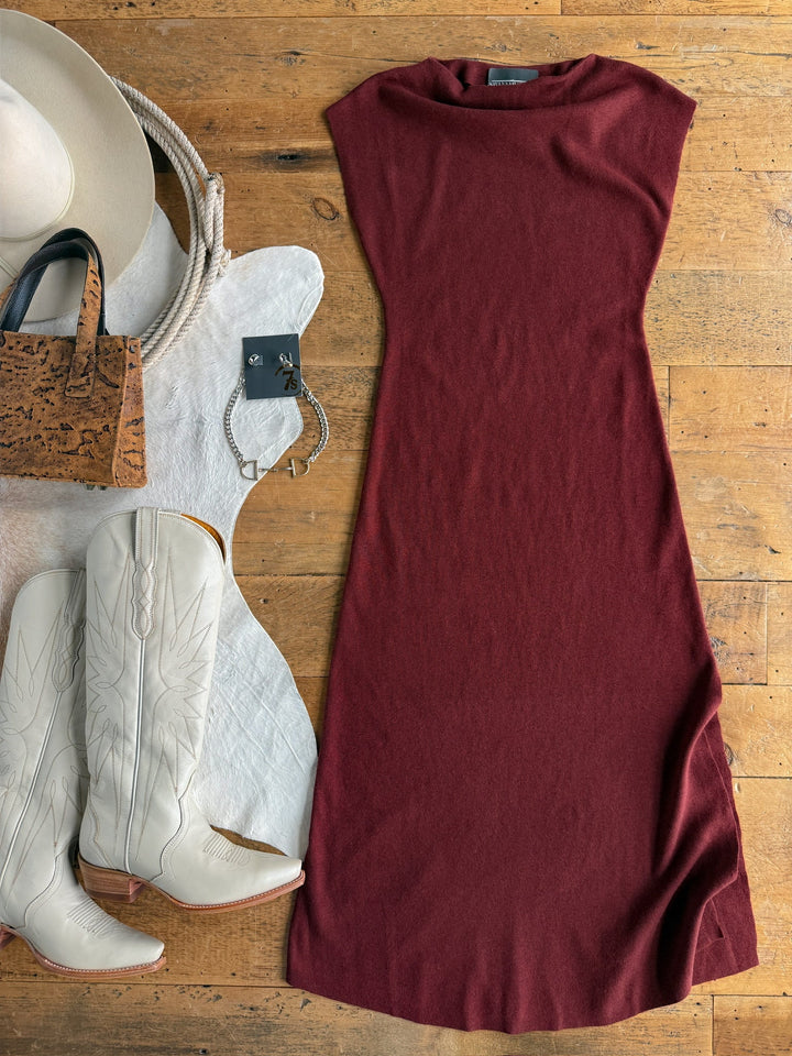 The Angola Wine Knit Dress by Savannah Sevens western life{&}style is styled with cream cowboy boots, a wide-brimmed hat, a cork handbag, a silver bracelet, and a black ring, all arranged on a wooden floor with a white fur rug.