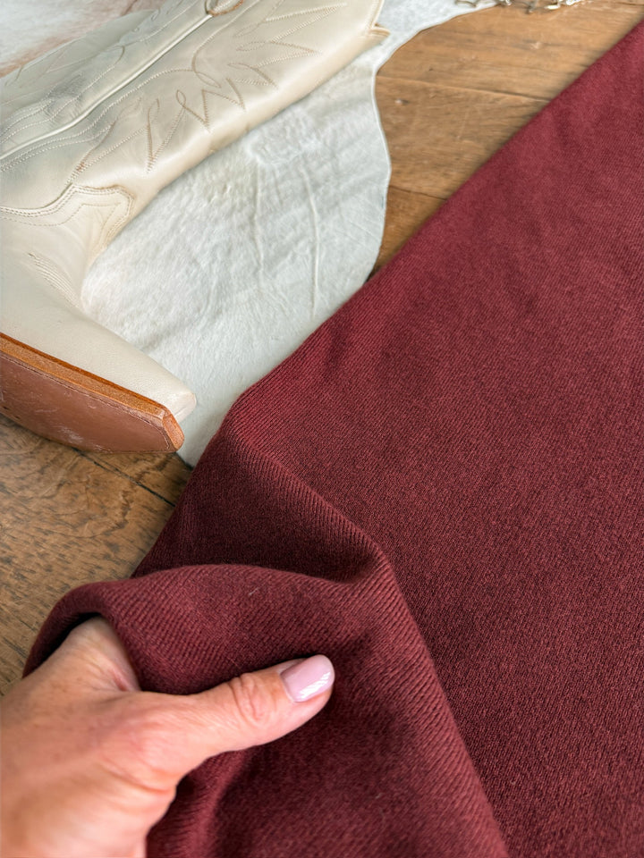 A hand with light pink nails holds burgundy fabric on a wooden surface beside a cream cowboy boot and a piece of white cloth, capturing the rich tones of the Angola Wine Knit Dress by Savannah Sevens western life{&}style.