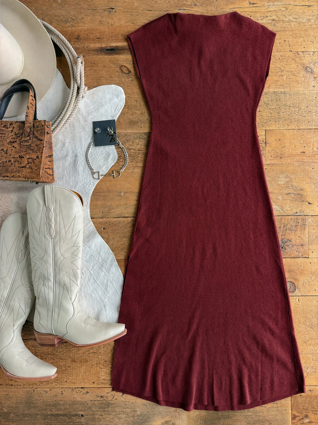 The Angola Wine Knit Dress from Savannah Sevens western life{&}style is laid out on a wooden floor, surrounded by white cowboy boots, a white hat, a cork handbag, a silver bracelet, and a pair of silver earrings.