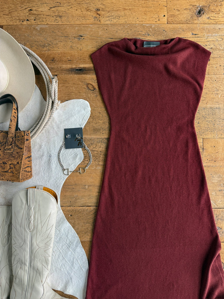 Flat lay featuring the Angola Wine Knit Dress by Savannah Sevens western life{&}style, paired with white cowboy boots, a tan hat, a cork handbag, a braided bracelet, and displayed on a white cowhide rug atop a wood floor.