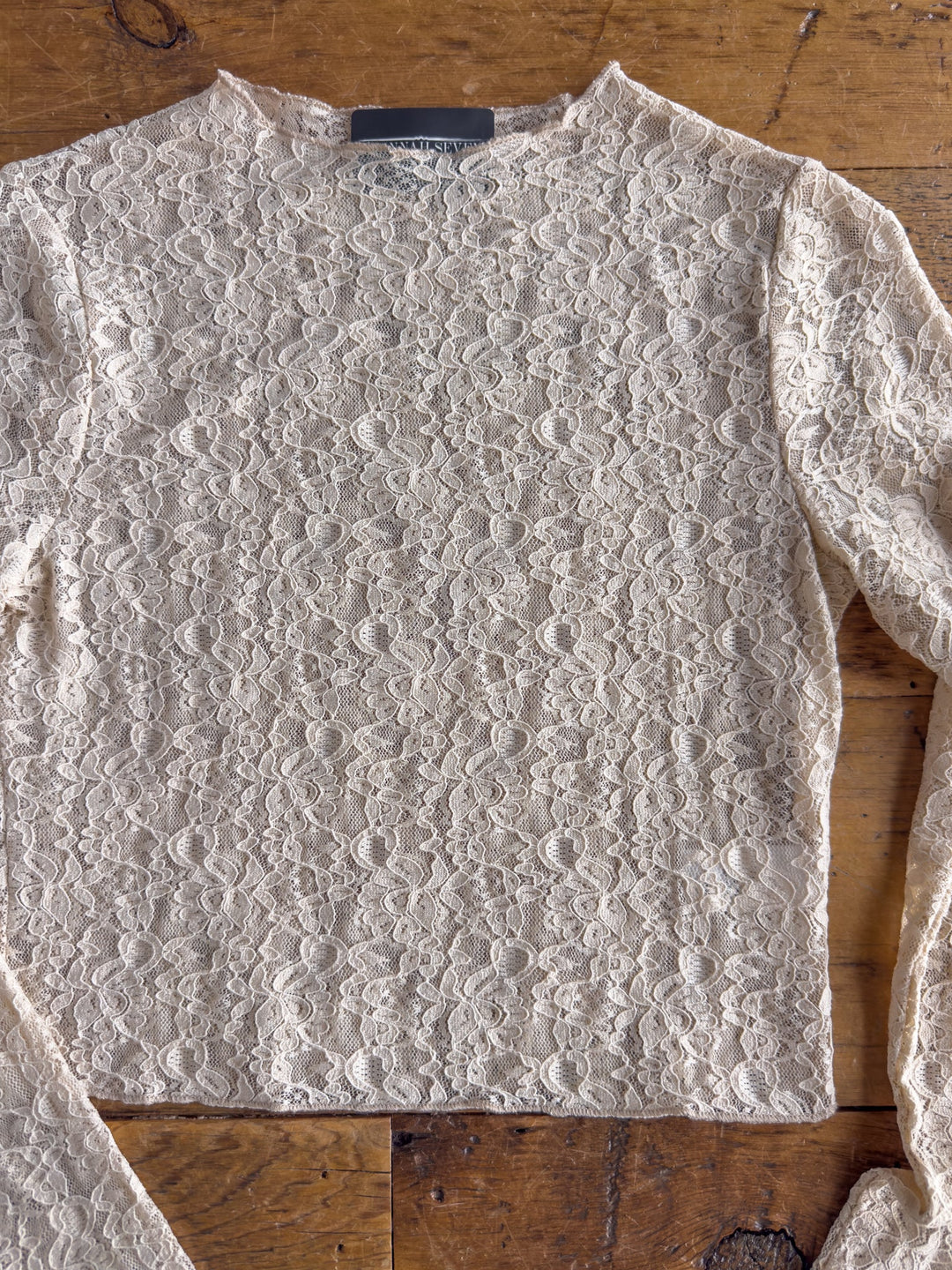 The Anniston Natural Lace Crop Top by Savannah Sevens western life{&}style, featuring cream-colored sheer mesh lace with a floral pattern and long sleeves, is laid flat on a wooden surface.