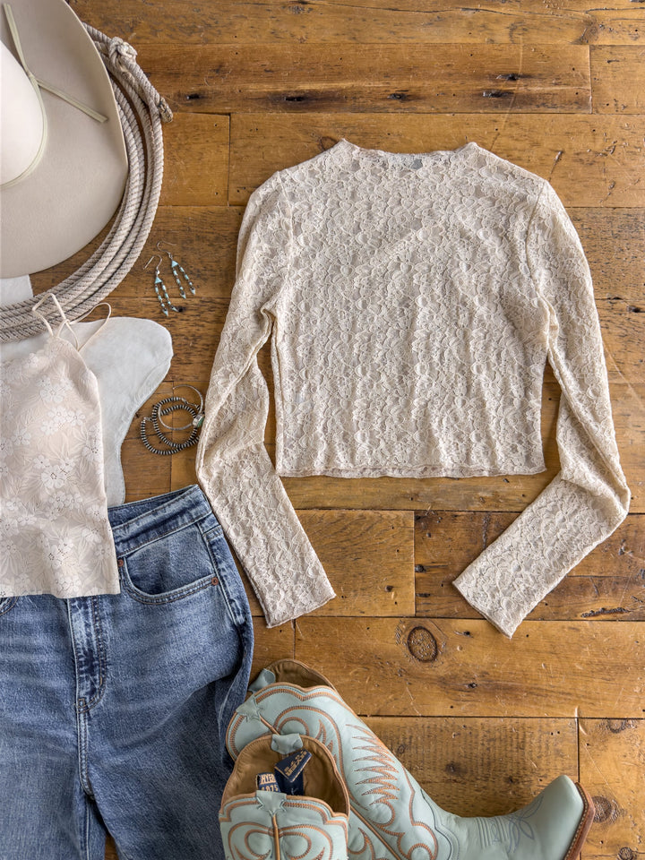 The Anniston Natural Lace Crop Top from Savannah Sevens western life{&}style, blue jeans, blue cowboy boots, a white cowboy hat, silver jewelry, and a matching lace bag are arranged on a wooden floor.