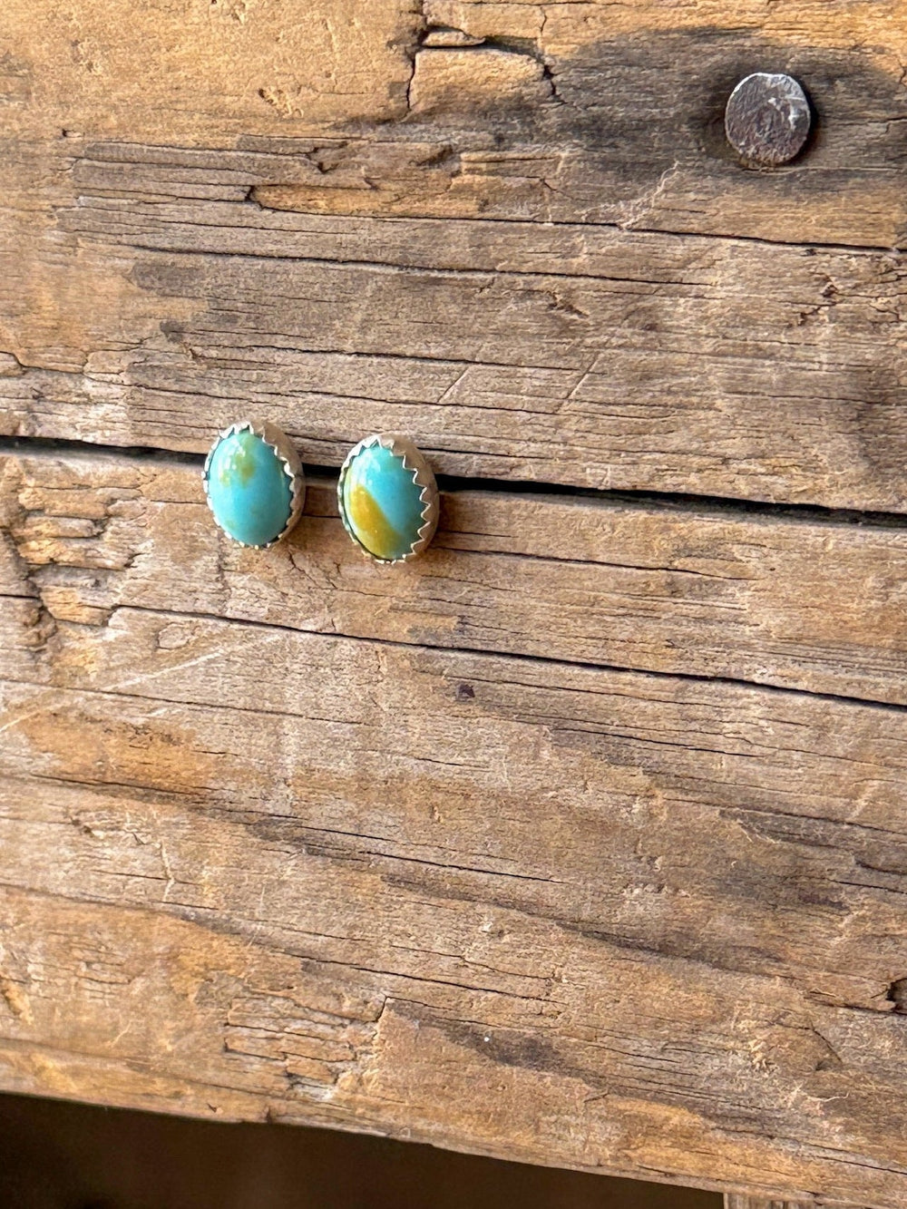 Anoka Turquoise Stud Earrings by Savannah Sevens western life{&}style, featuring vibrant green oval stones set in sterling silver, are displayed on a rustic, weathered wooden surface with visible grain and a metal nail.