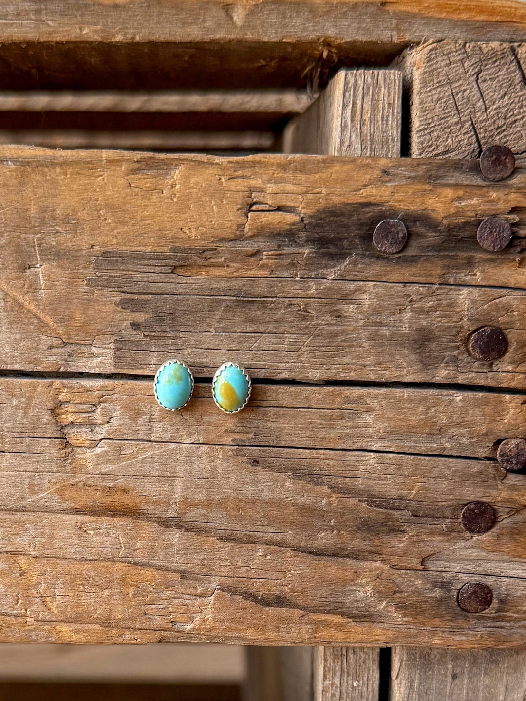 A pair of Anoka Turquoise Stud Earrings by Savannah Sevens western life{&}style, featuring round Navajo turquoise set in sterling silver, rests on a rustic wooden surface with visible nails and weathered textures.